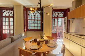 Ein Essbereich mit einem runden Tisch, Stühlen und einer Couch. Es gibt Fenster mit verzierten Glasscheiben und eine Küchenzeile mit Holzausbau.