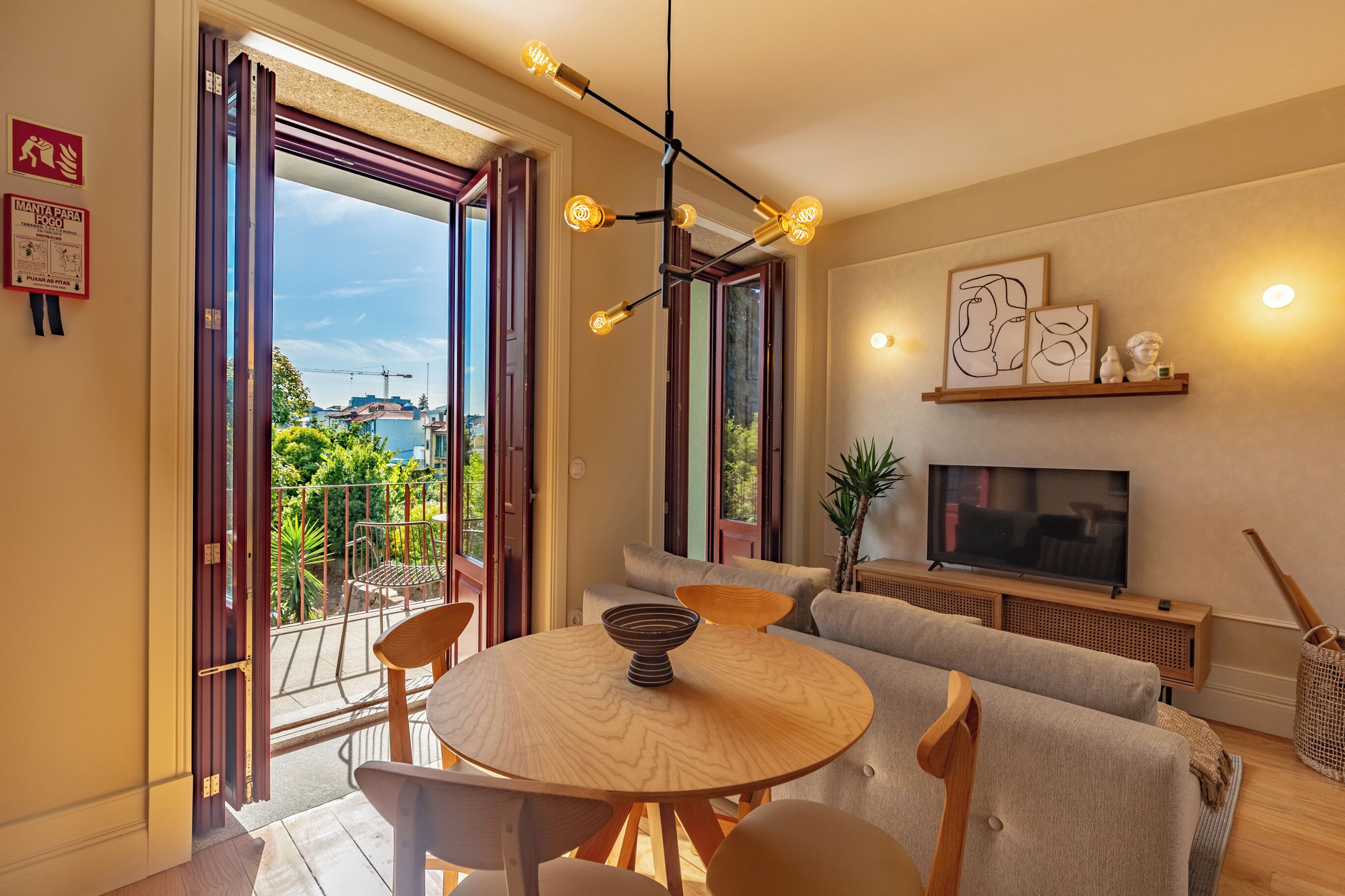 A modern living room with a round wooden table, a sofa, a TV, and a balcony with a view.