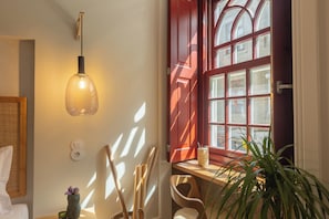 Ein Zimmer mit einem Bett, einem Nachttisch mit Lampe, einem Fenster mit roten Lamellen und einer Zimmerpflanze.