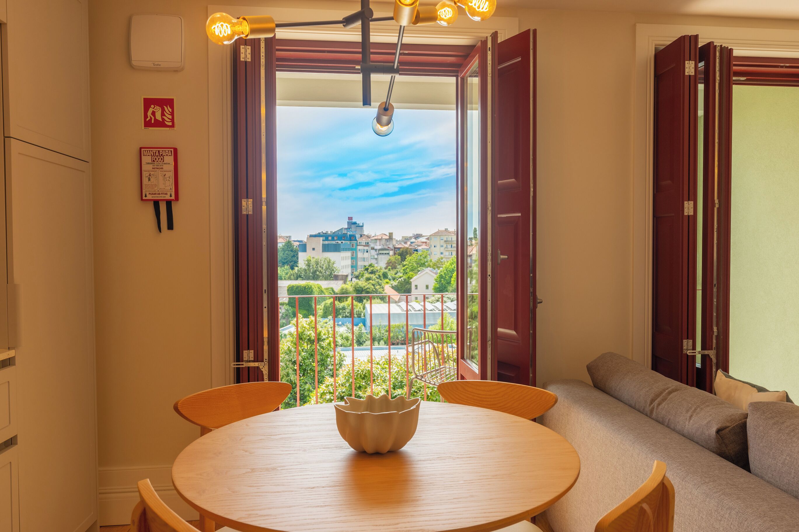 Ein Zimmer mit einem runden Holztisch, einer Couch und Blick auf Gebäude und Grünflächen durch offene Türen.