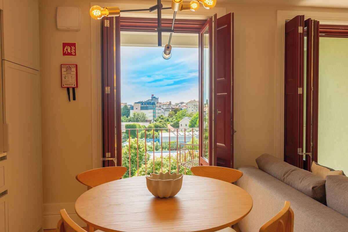 Quarto com uma mesa de madeira redonda, um sofá e vista para prédios e vegetação através de portas abertas.