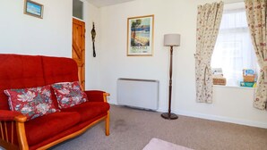 A living room with a red sofa, a radiator, a lamp, a window with curtains, and a framed picture on the wall. - Chyancrowse (Penzance)