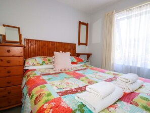 Ein Schlafzimmer mit einem hölzernen Kopfende, einem farbenfrohen Bettbezug, einer Holzkommode und einem Fenster mit durchscheinenden Vorhängen.