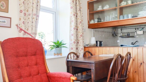 A dining area with a red upholstered chair, wooden table, and chairs, a window with floral curtains, and a kitchen with wooden cabinets and glass-fronted shelves. - Chyancrowse (Penzance)