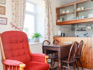 A dining area with a red upholstered chair, wooden table, and chairs, a window with floral curtains, and a kitchen with wooden cabinets and glass-fronted shelves.
