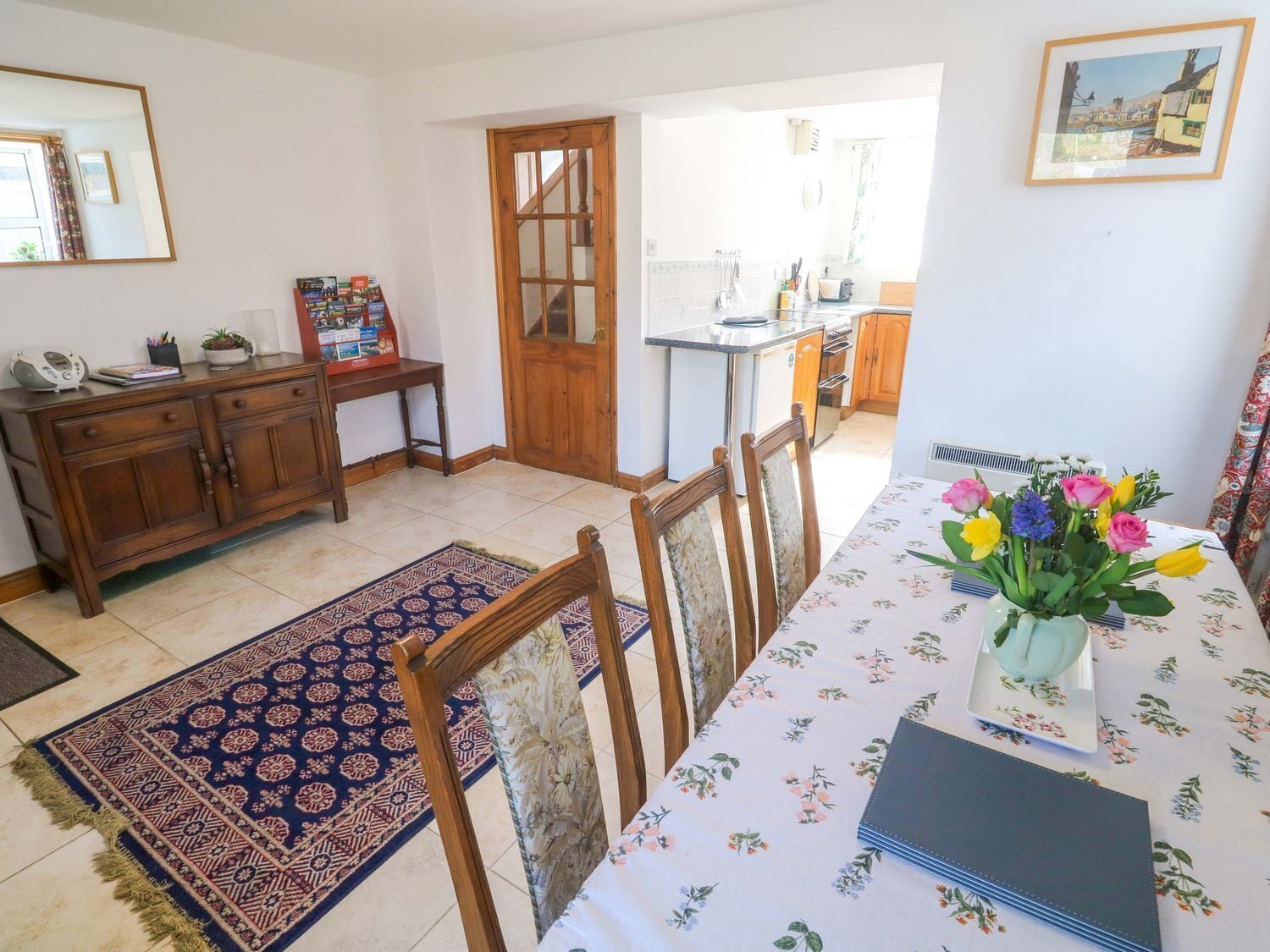 Un comedor con una mesa dispuesta para cuatro, un armario de madera, una alfombra y una cocina al fondo.