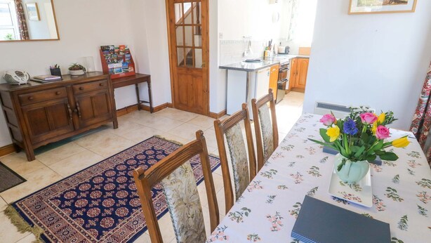 Un comedor con una mesa dispuesta para cuatro, un armario de madera, una alfombra y una cocina al fondo.