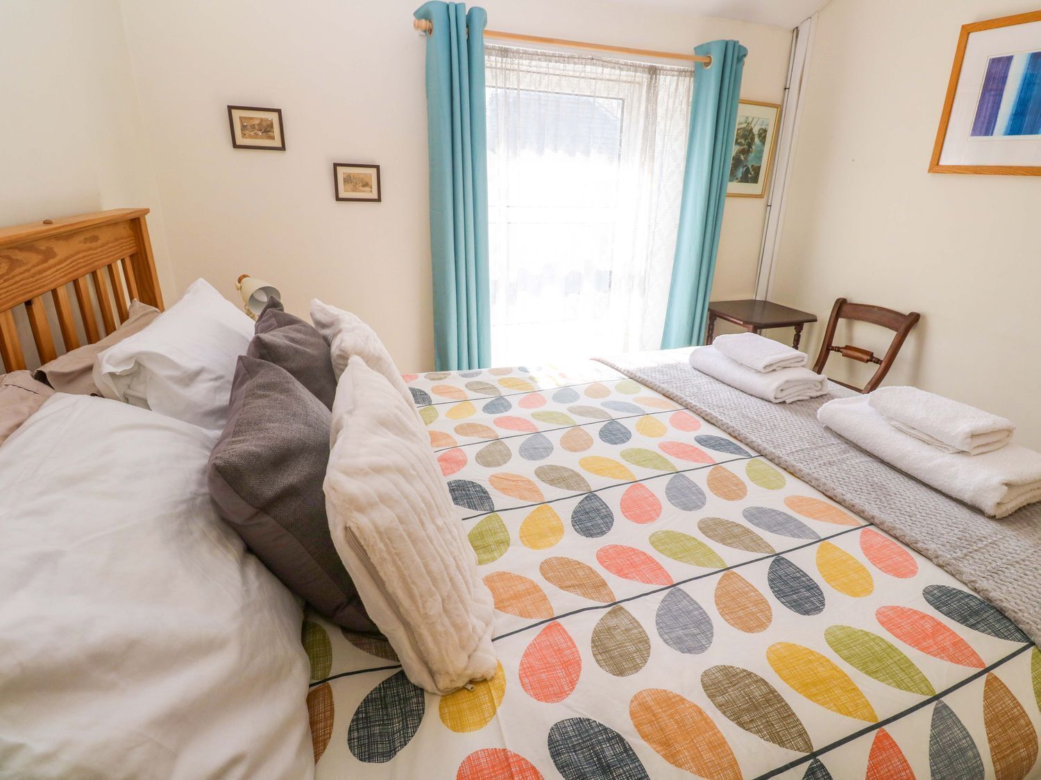 Un dormitorio con cama, almohadas, una mesita de noche, una silla y una ventana con cortinas.