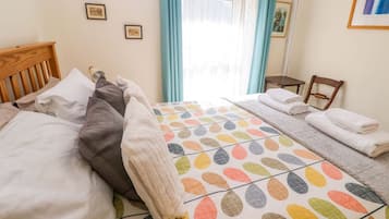 Un dormitorio con cama, almohadas, una mesita de noche, una silla y una ventana con cortinas.