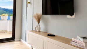 Un salon moderne avec un téléviseur à écran plat fixé au mur, une console en bois et une porte-fenêtre coulissante donnant sur un balcon avec vue sur les montagnes.