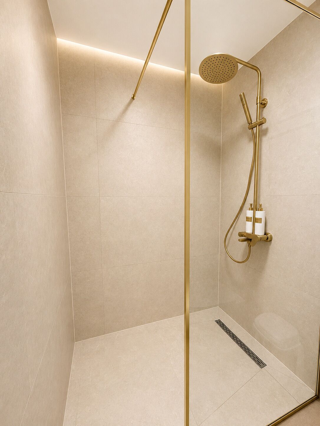 En modern dusch med ett guldfärgat duschmunstycke och vred, beige kakel på väggarna och en glasvägg.