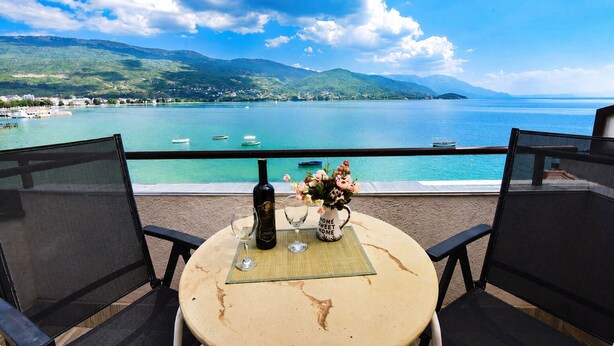 Une table avec une bouteille et des verres, donnant sur un lac où l’on aperçoit des bateaux et des montagnes.