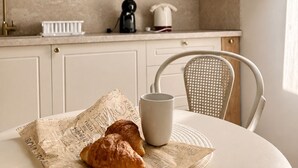 Une cuisine avec une lucarne, une table blanche sur laquelle se trouvent une tasse et des croissants, et une chaise en osier.