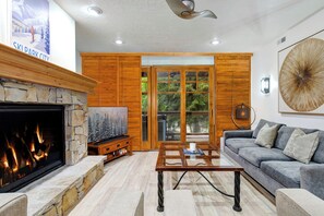 A living room with a stone fireplace, a television, a large window with a view of trees, and a modern ceiling fan. - Managed by Park City Lodging-Prime Location Steps from Skiing, Dining/Shopping (Park City)