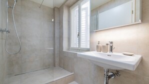 Un bany modern amb un box de dutxa de vidre, un lavabo blanc amb un mirall a sobre i una finestra amb persianes.