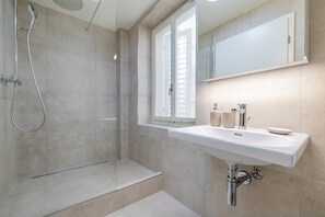 Ein modernes Badezimmer mit einer Duschkabine aus Glas, einem weißen Waschbecken mit Spiegel darüber und einem Fenster mit Jalousien.