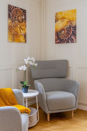 Ein Zimmer mit einem grauen Sessel, einem kleinen weißen Tisch, einer Topf-Orchidee und zwei abstrakten Bildern an der Wand.