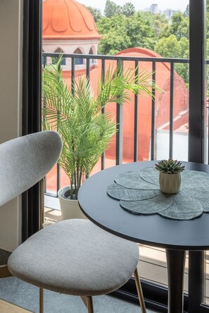 Un balcon avec une table ronde, deux chaises, un pot de fleurs et une vue sur un immeuble avec un toit rouge.
