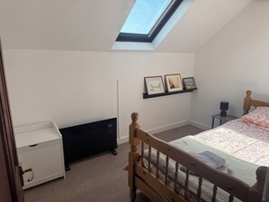 A bedroom with a skylight, a bed with a patterned bedspread, a bedside table with a lamp, and a shelf with framed pictures. - Penpegws (Aberystwyth)
