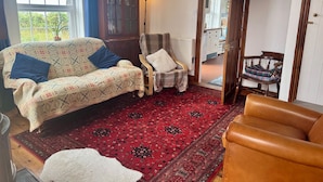 Un salón con un sofá estampado, una silla de madera, una alfombra roja y vistas a través de un ventanal abierto.