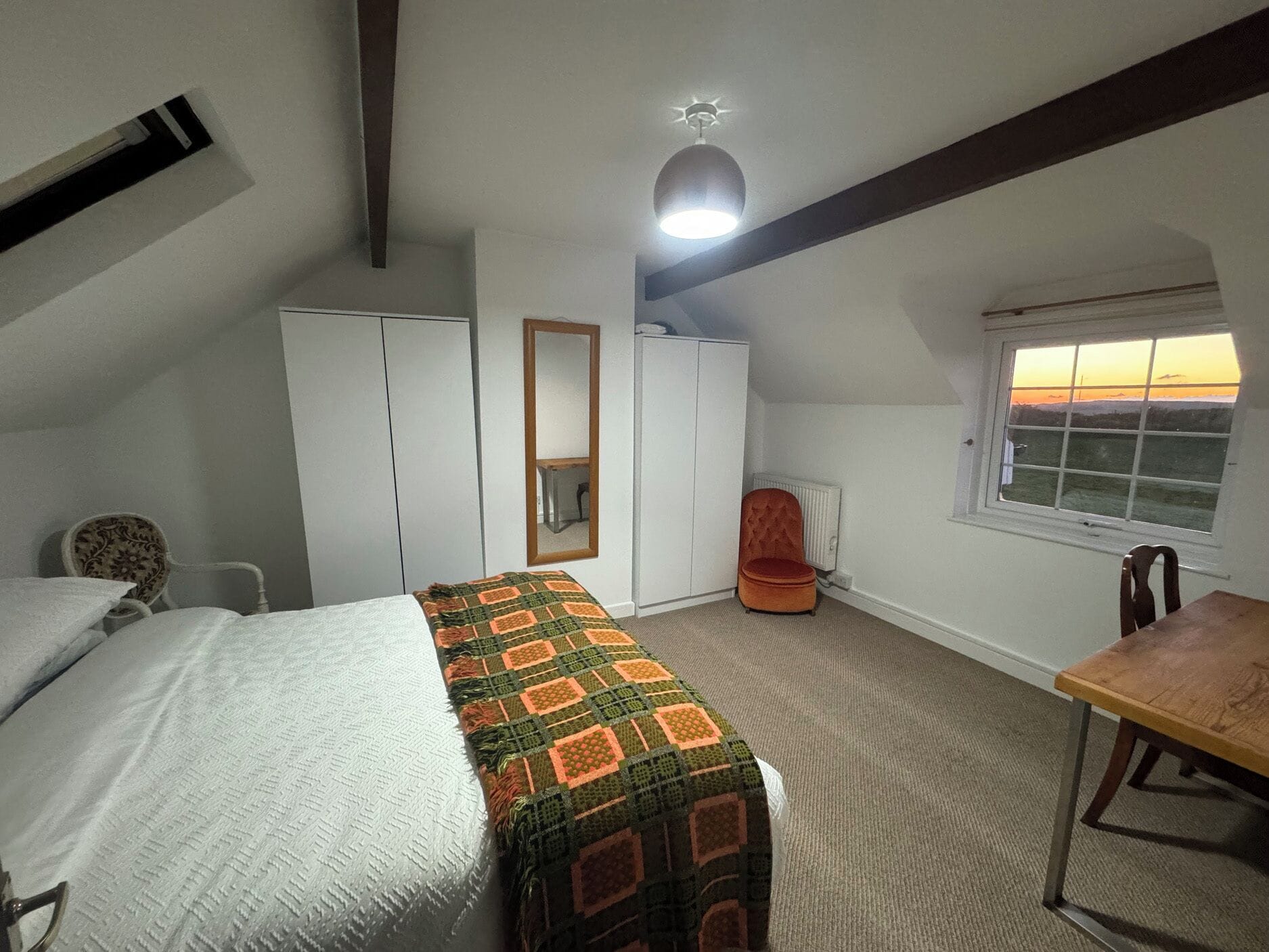 Una habitación con cama, escritorio, silla, armario, radiador y ventana con vistas.