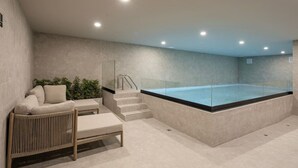 Un espace spa moderne doté d’un jacuzzi entouré de parois de verre, d’un coin salon avec un canapé et un pouf, et d’un escalier menant au jacuzzi.