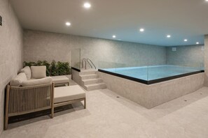 Un espace spa moderne doté d’un jacuzzi entouré de parois de verre, d’un coin salon avec un canapé et un pouf, et d’un escalier menant au jacuzzi.