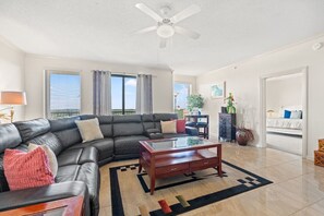A living room with a black leather sofa, a wooden coffee table, and a view of the outdoors through large windows. - 9th-Floor Ocean View Condo, Pool & Hot Tub (Myrtle Beach)
