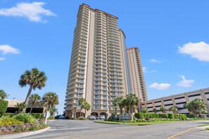 A tall, beige multi-story building with balconies, surrounded by palm trees and a parking lot. - 9th-Floor Ocean View Condo, Pool & Hot Tub (Myrtle Beach)
