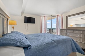Ein Schlafzimmer mit Bett, einem an der Wand montierten Fernseher, einem Balkon mit Vorhängen und einer Kommode.