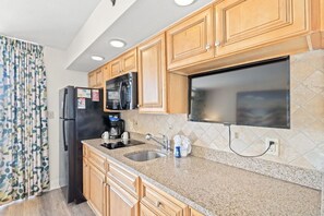A kitchen with wooden cabinets, a refrigerator, a microwave, a coffee maker, and a large window. - Oceanfront Resort with Lazy River (North Myrtle Beach)