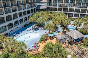 A hotel with a central pool surrounded by palm trees, a separate pool area, and a building with multiple balconies. - Oceanfront Resort with Lazy River (North Myrtle Beach)
