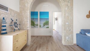 Una habitación con un arco de piedra, suelo de madera, un sofá azul, un aparador de madera y dos cuadros con escenas de playa.