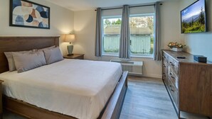 Un dormitorio con cama, mesita de noche, cómoda, ventana con cortinas y televisor instalado en la pared.