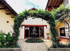 Une cour avec une fontaine en pierre, une arche couverte de plantes grimpantes et l’entrée d’un bâtiment surmontée d’un toit rouge.
