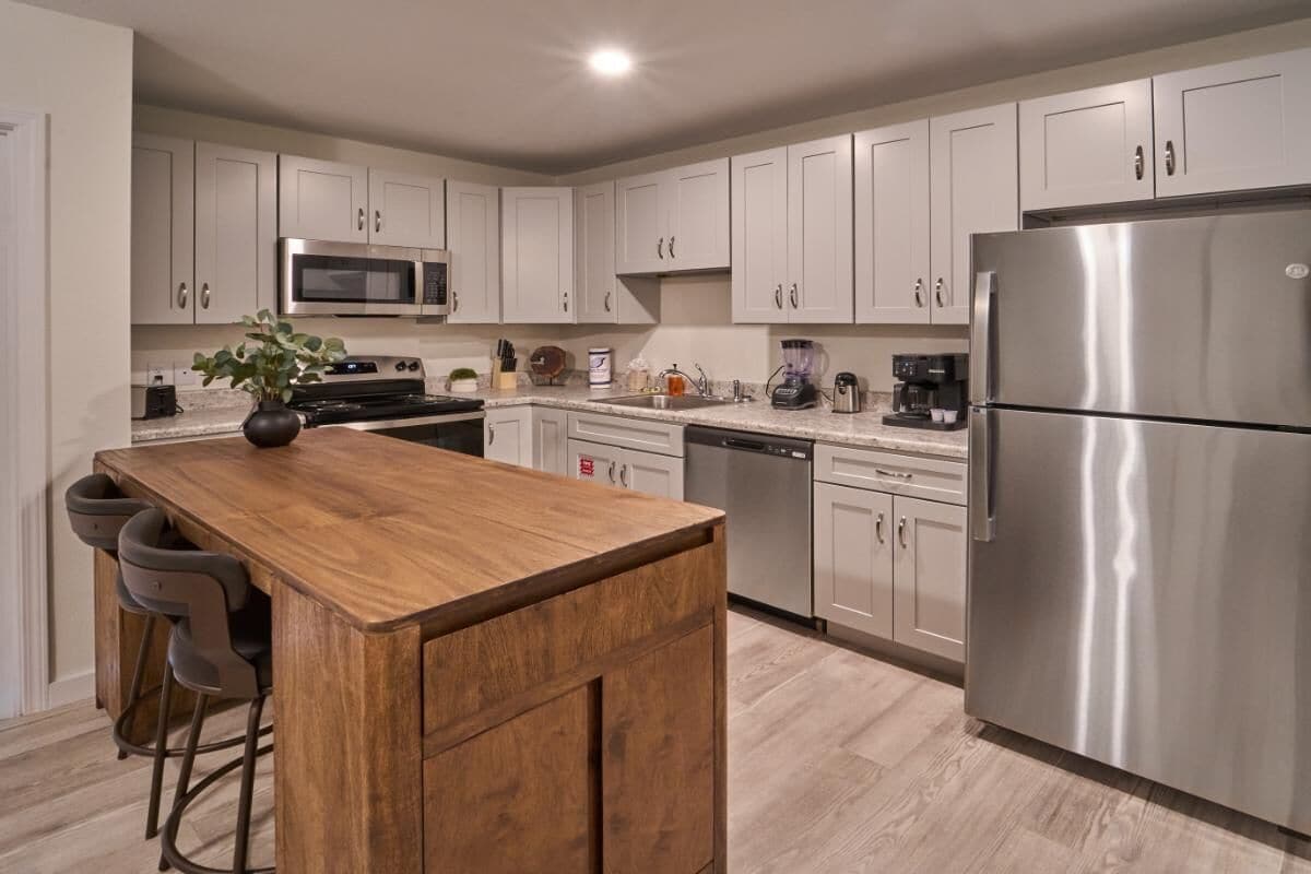 A modern kitchen with white cabinets, stainless steel appliances, a wooden island, bar stools, and a built-in dishwasher.