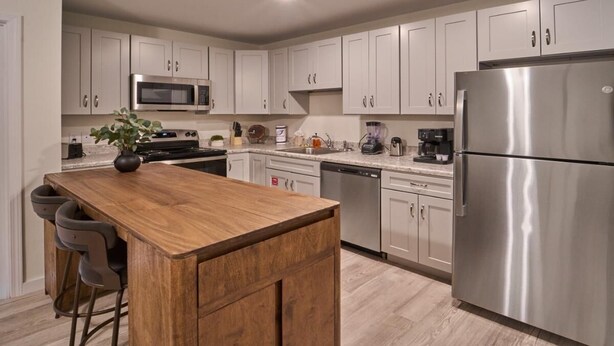 Une cuisine moderne avec des meubles blancs, des appareils en acier inoxydable, un îlot central en bois, des tabourets de bar et un lave-vaisselle encastré.