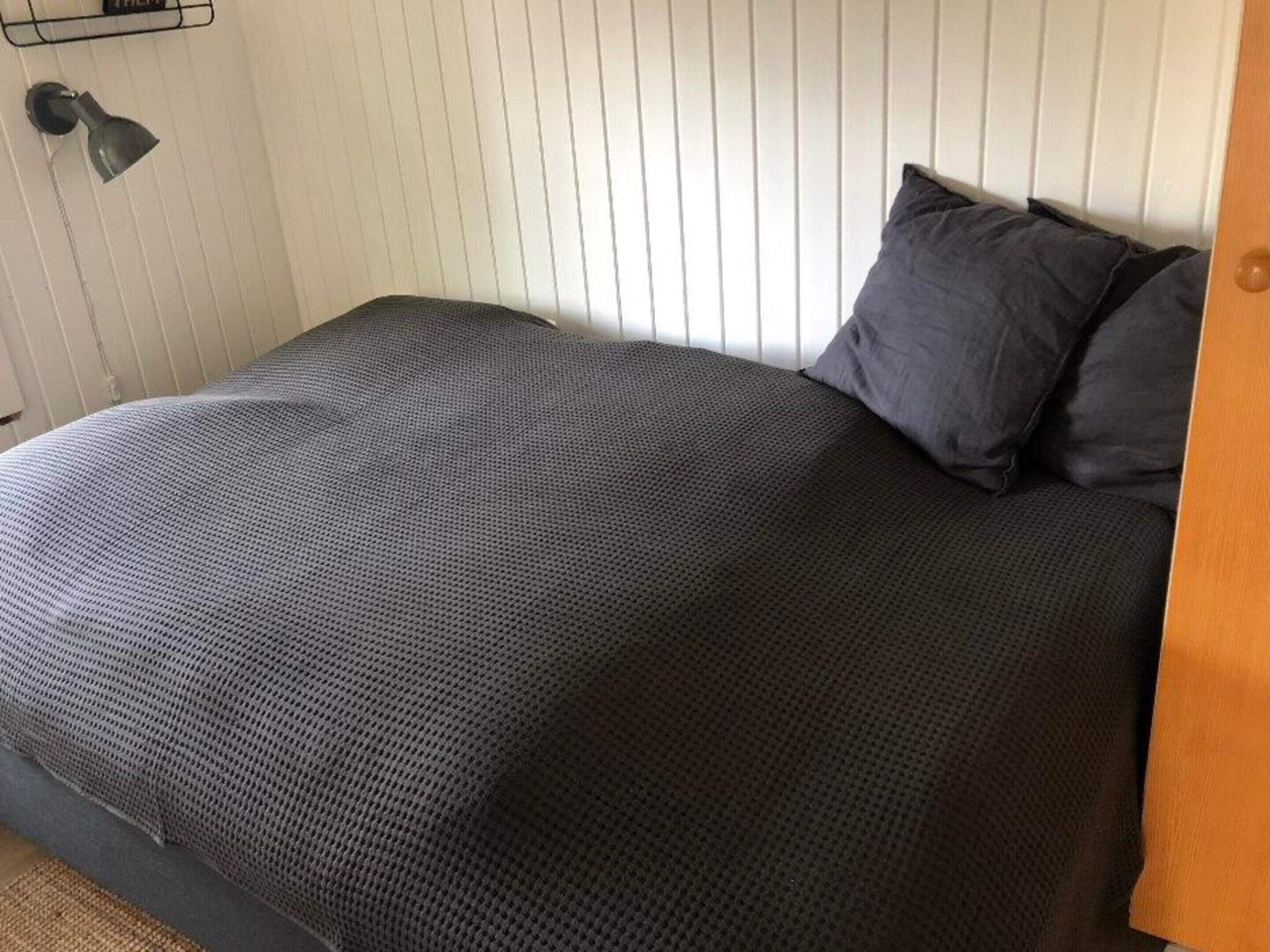 A bed with a dark patterned bedspread, a single dark pillow, and a wall-mounted lamp.
