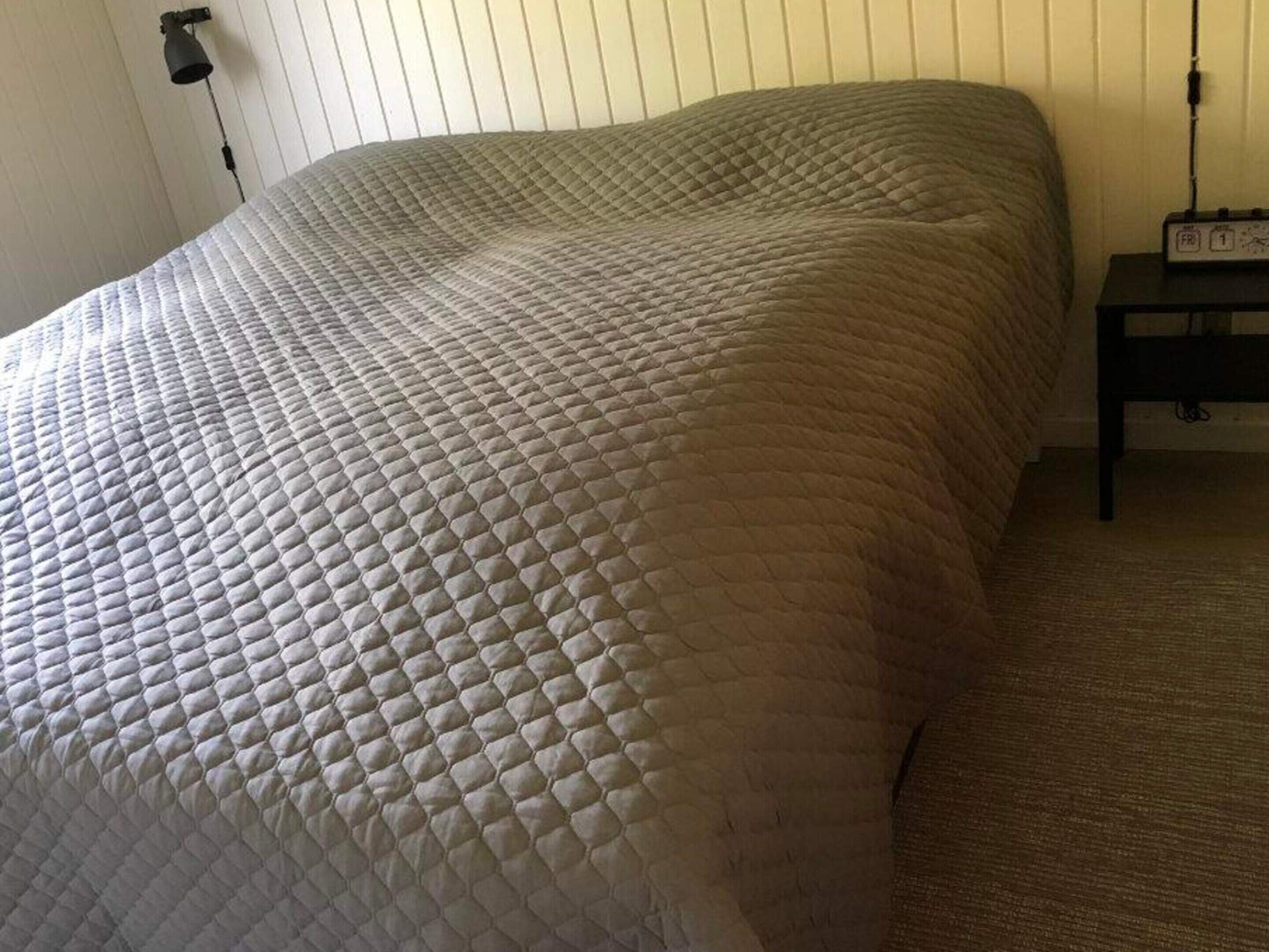 Een bed met een gewatteerde spreidek, een nachtkastje met een lamp en een wandcontactdoos.