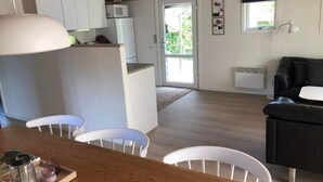 A modern kitchen with white cabinets, a dining area with a wooden table and white chairs, and a living area with a black sofa and a small white table. - 4 Star Holiday Home in Esbjerg V (Esbjerg)