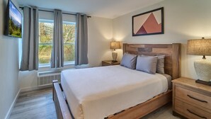 Un dormitorio con una cama, una mesita de noche, una lámpara, una ventana con cortinas y un cuadro colgado en la pared.