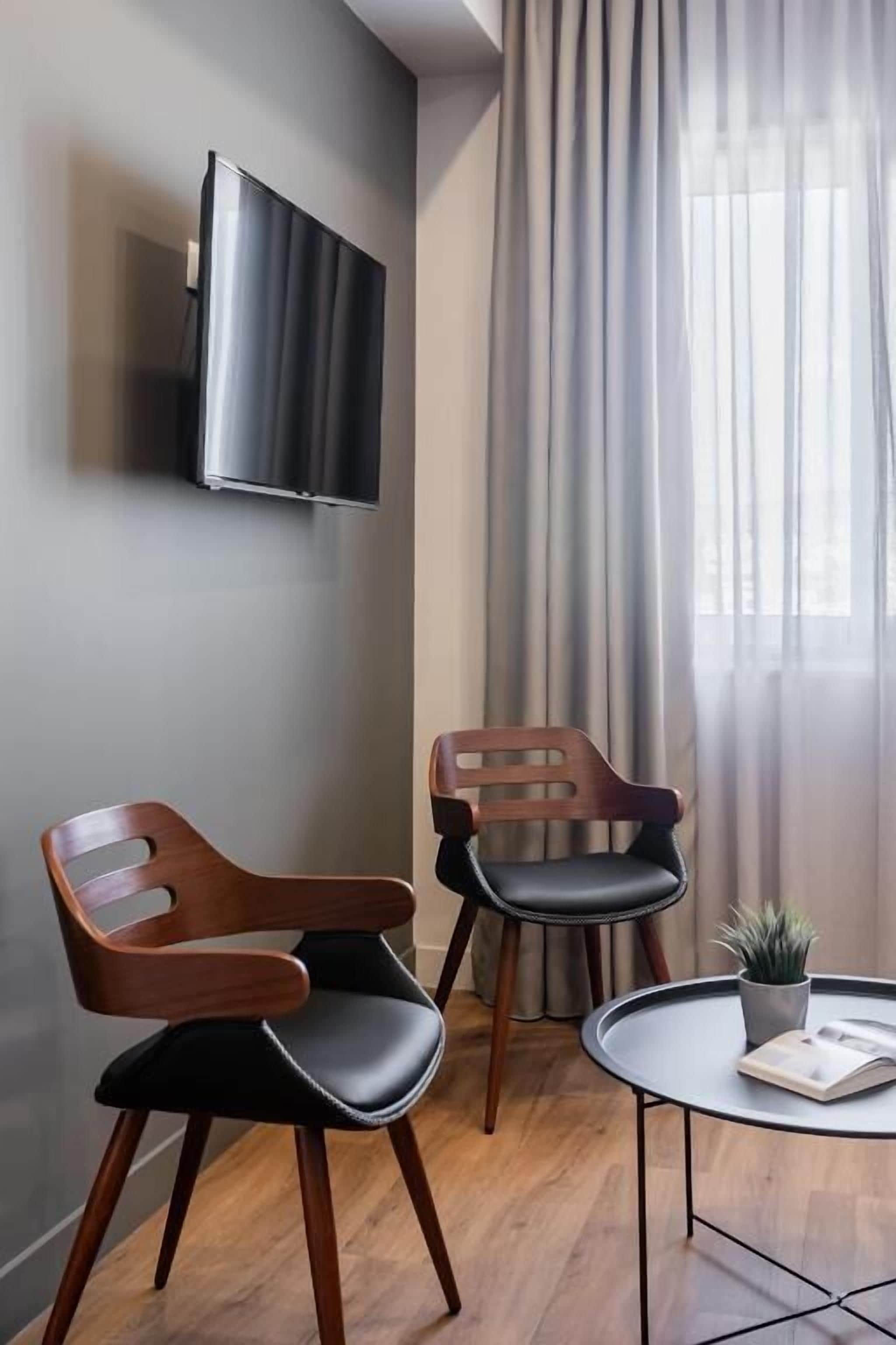 A modern room with a flat-screen TV mounted on the wall, two wooden chairs with black cushions, a small round table with a plant and a book, and a large window with sheer curtains.