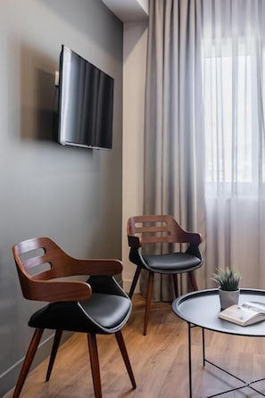 A modern room with a flat-screen TV mounted on the wall, two wooden chairs with black cushions, a small round table with a plant and a book, and a large window with sheer curtains. - Au Piree by Calon (Pireas)