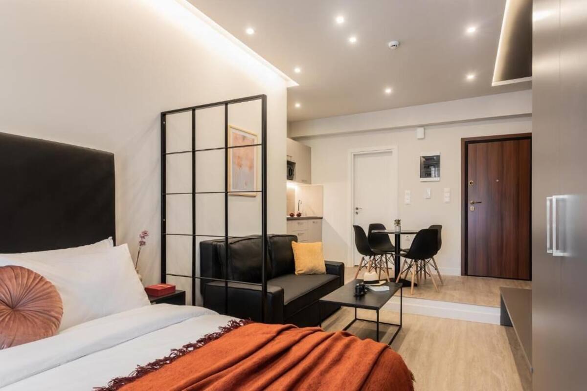 A modern hotel room with a bed, a sofa, a dining table with chairs, a kitchen area, and a separate door.