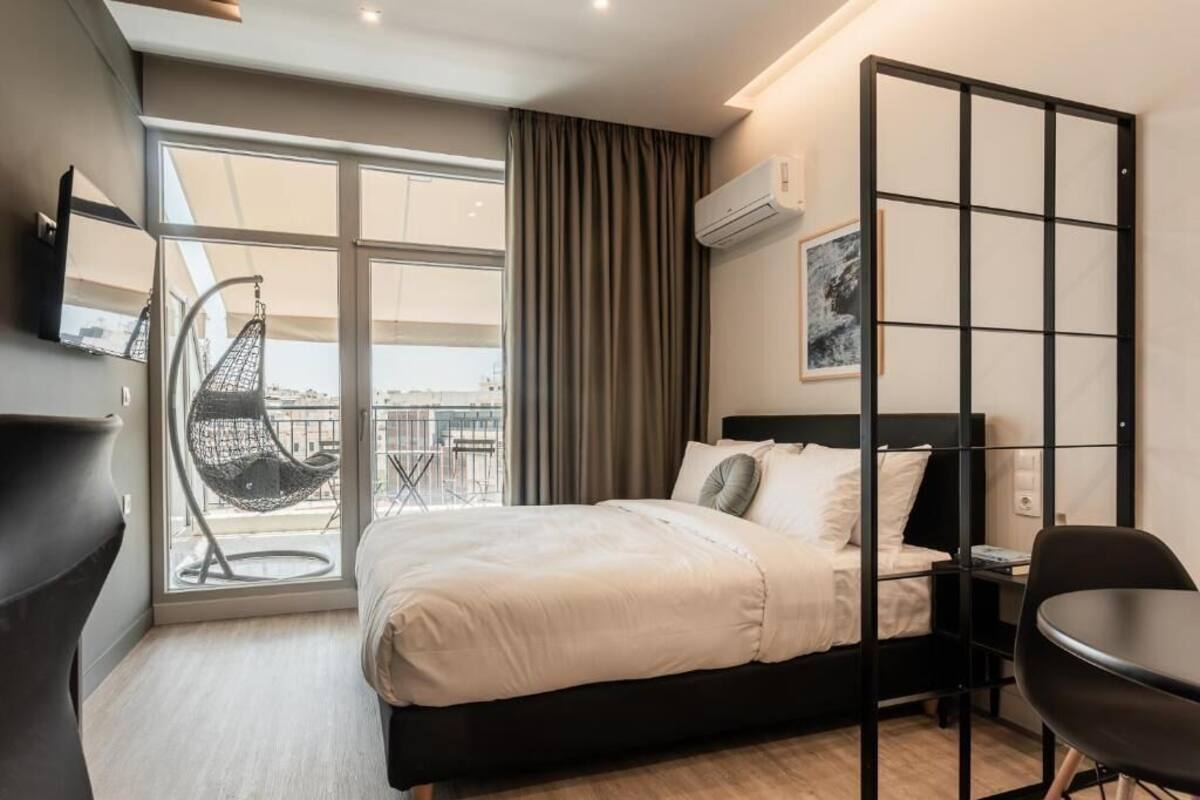 A modern hotel room with a bed, a chair, a desk, a large window, and a view of the city.