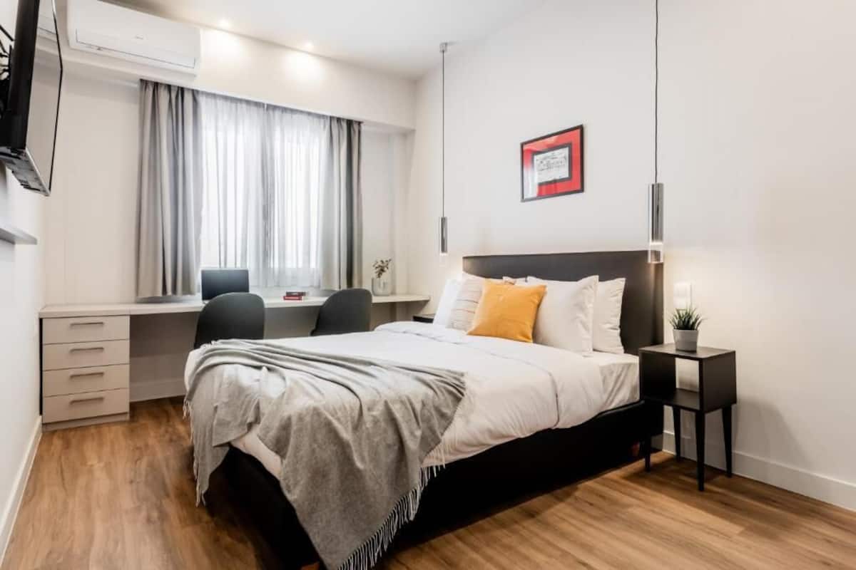 A bedroom with a bed, bedside table, desk, chair, and an air conditioning unit.