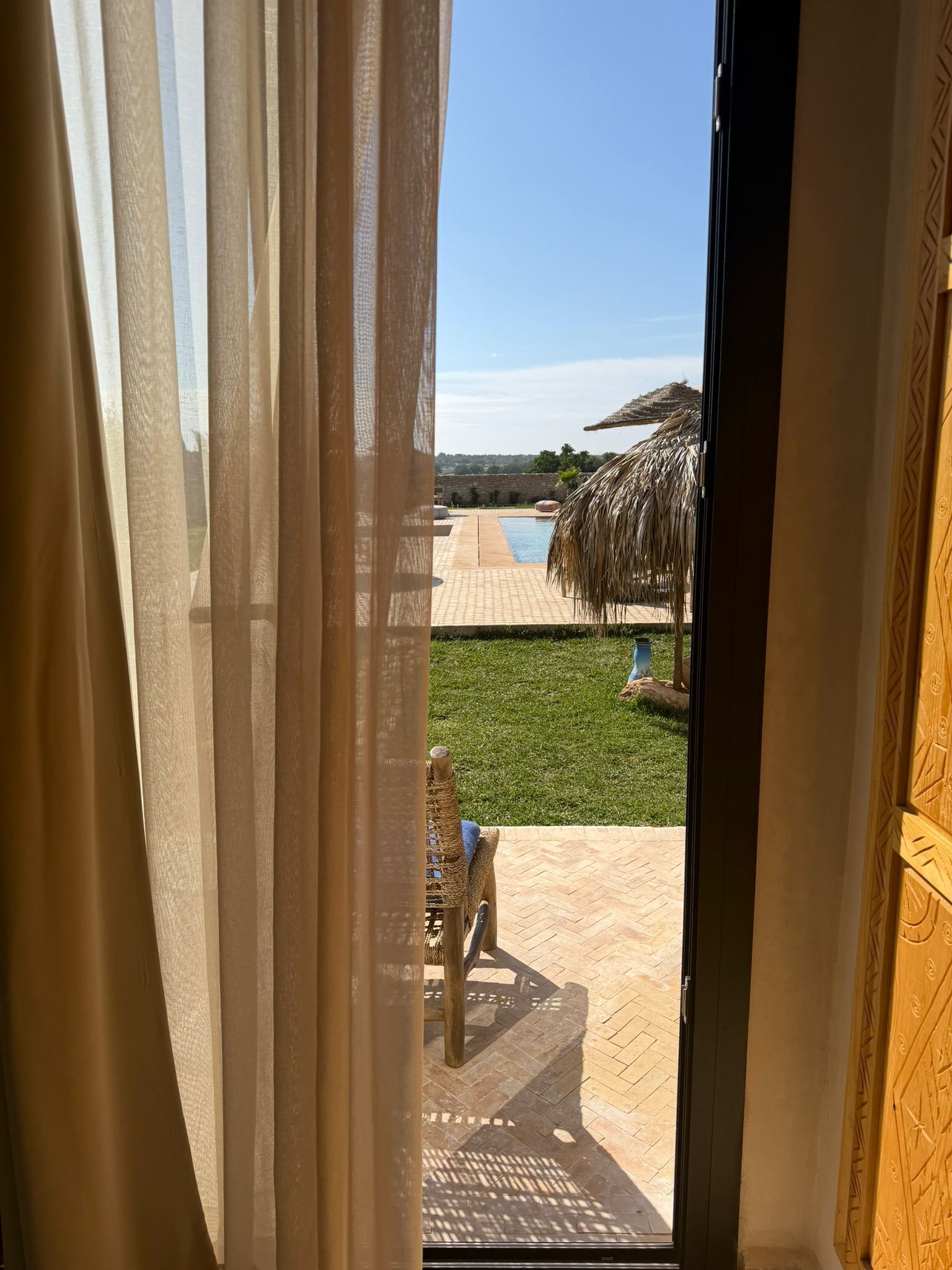 Blick durch ein Fenster mit durchsichtigen Vorhängen auf einen Pool, eine Rasenfläche und ein Gebäude mit Strohdach.
