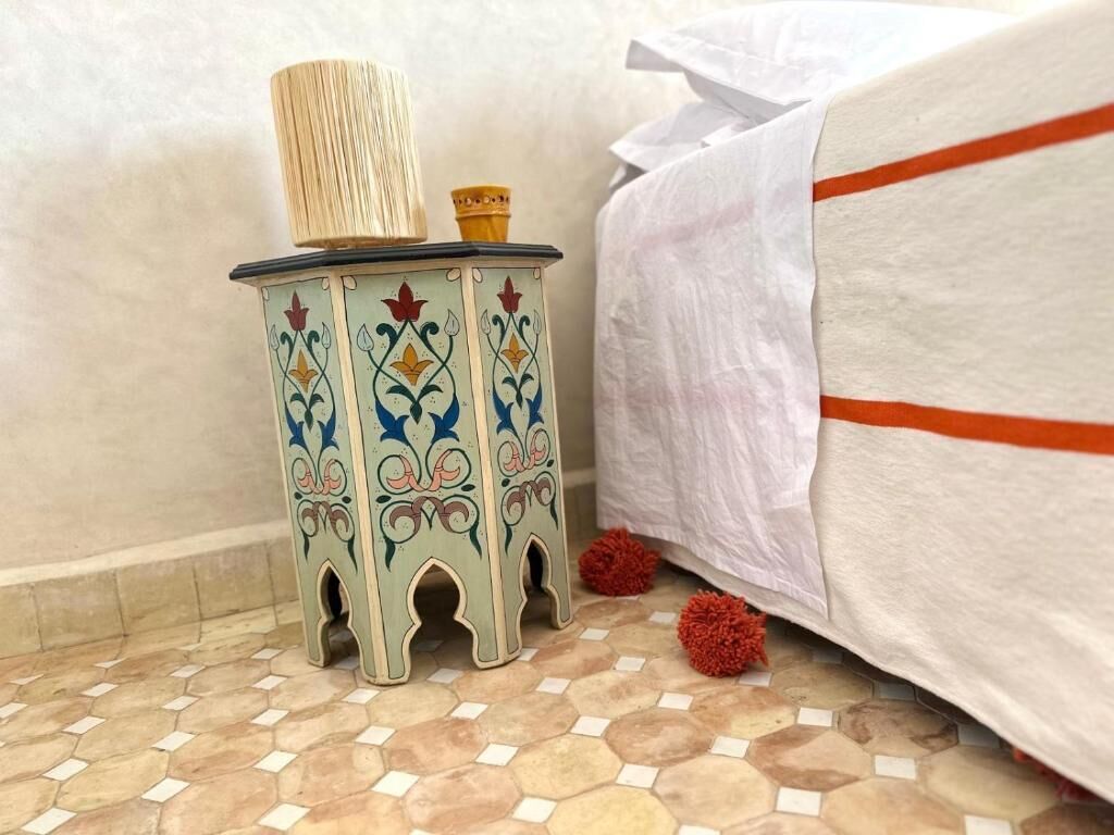 A small, decorative stool with a woven top and a small container beside a bed with a white cover and orange trim.