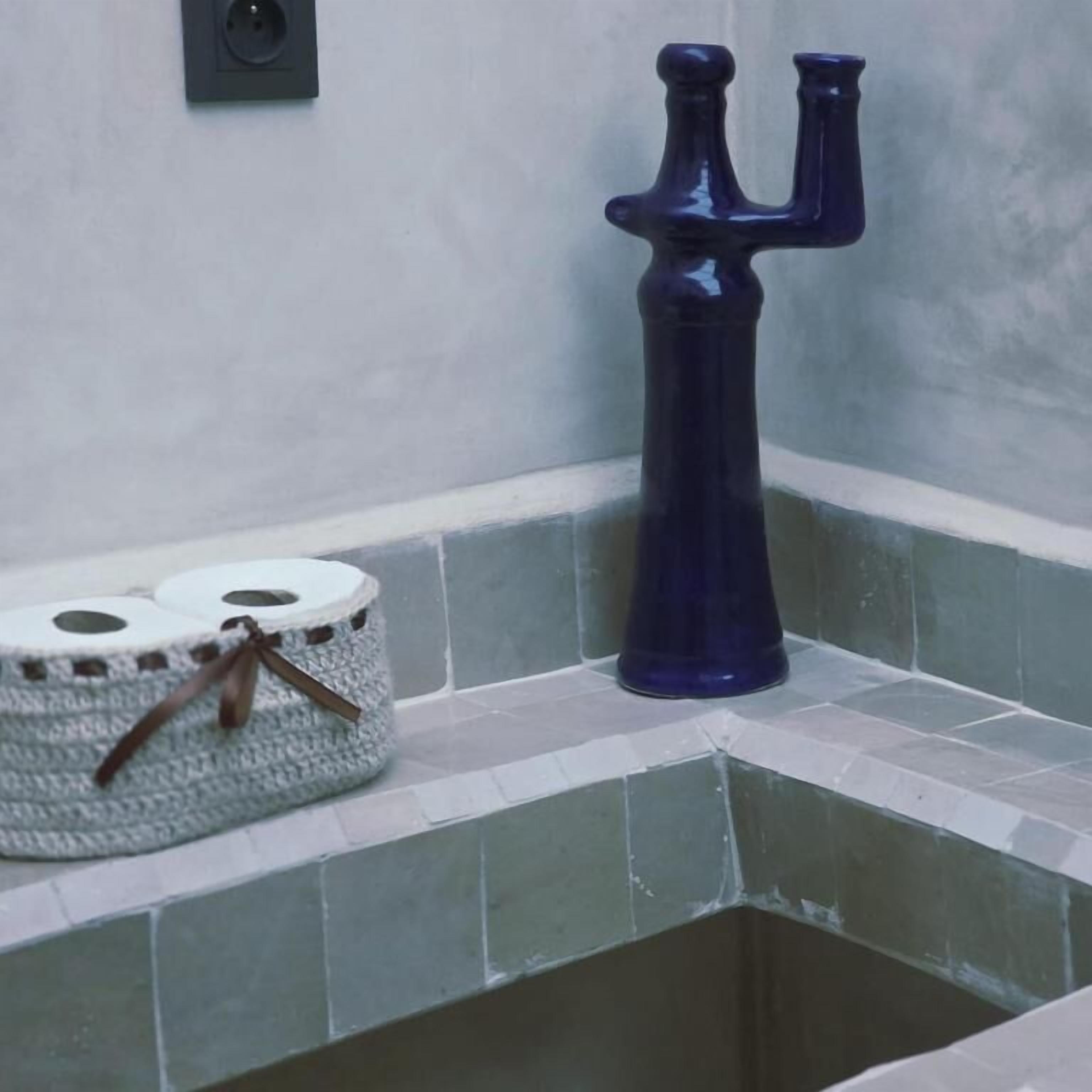 A bathroom with a blue vase, a basket of toilet paper, and a wall-mounted light fixture.
