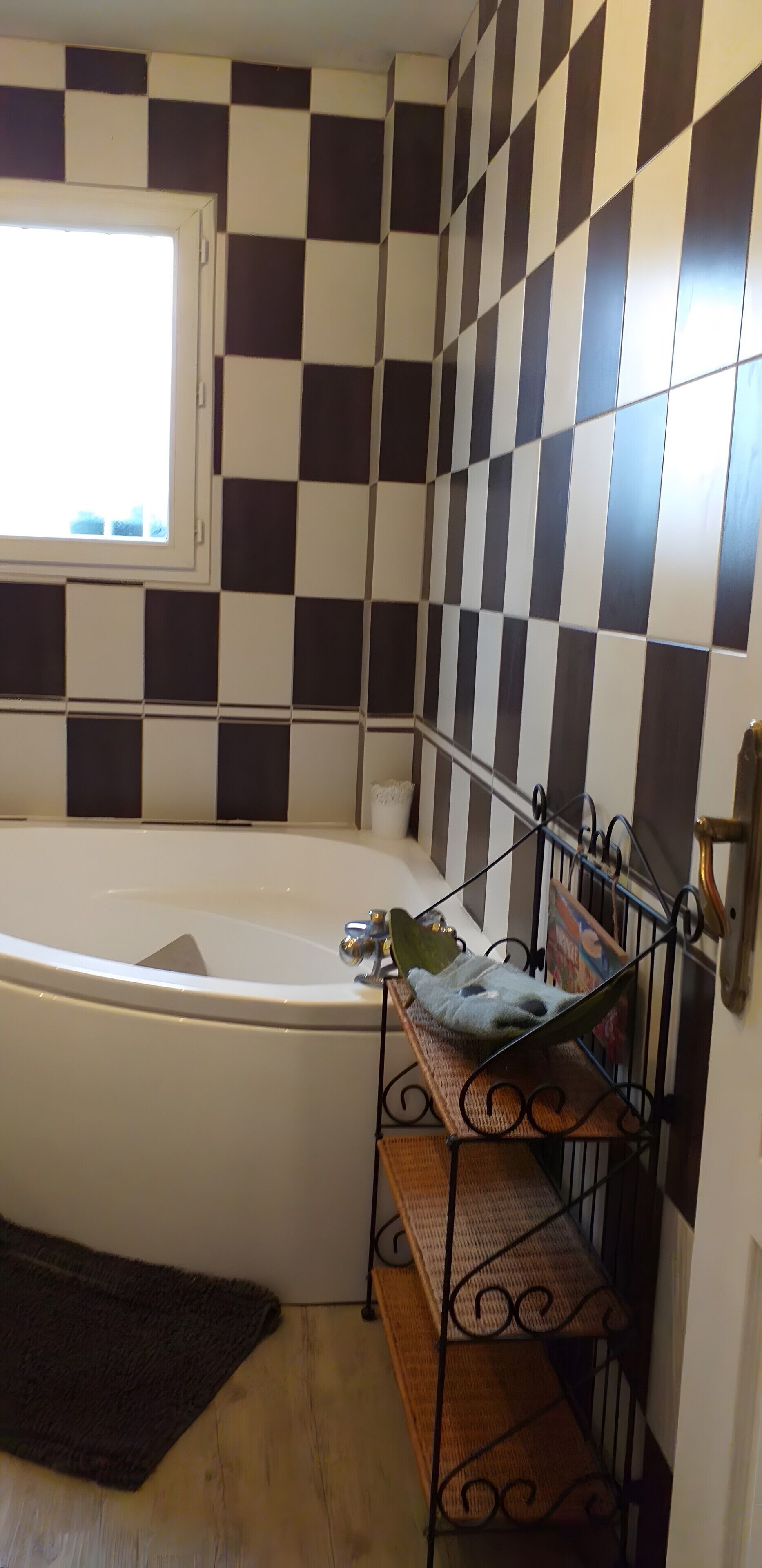 A bathroom with a checkered tile wall, a bathtub, a window, and a metal shelf with a towel.